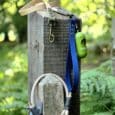 Blue BioThane dog lead and green waste bag dispenser attached to wooden post in garden setting.