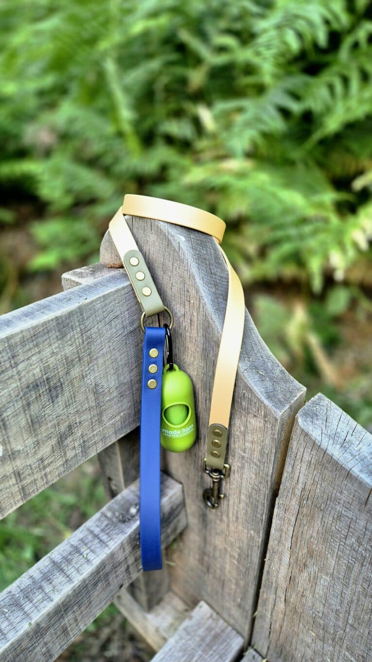 Blue BioThane dog lead with cream handle and green waste bag dispenser attached to wooden fence.