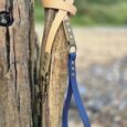 Handmade BioThane dog lead with cream and blue straps attached to weathered wooden post by beach.