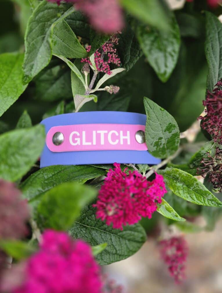 Pink nameplate engraved with 'GLITCH' on a blue and pink handmade BioThane dog collar among pink flowers.