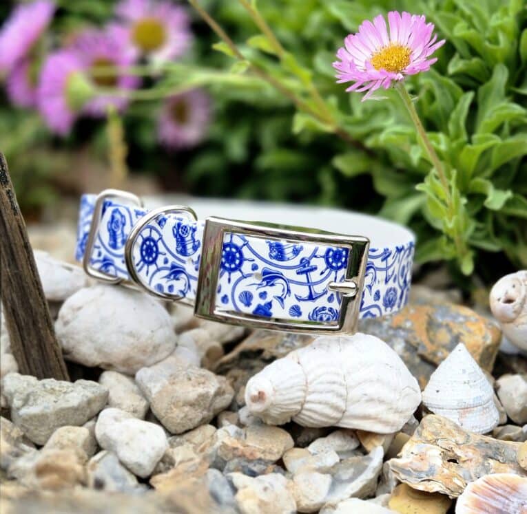 Handmade BioThane dog collar with blue and white nautical print design, displayed on pebbles with flowers.