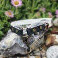 Black handmade BioThane dog collar with brass fittings displayed on rocks against purple flowers.