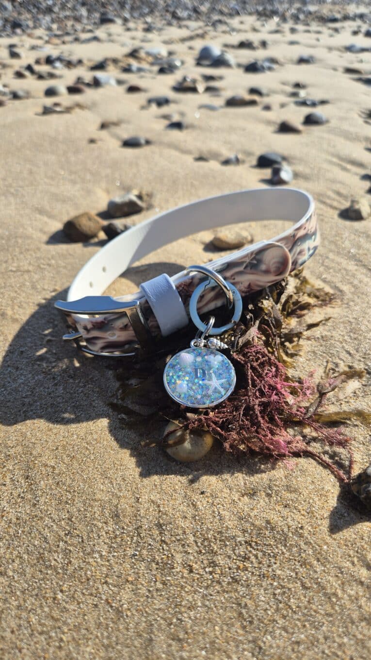 Coastal-printed BioThane collar with matching personalised resin tag on sandy beach amongst seaweed and pebbles.