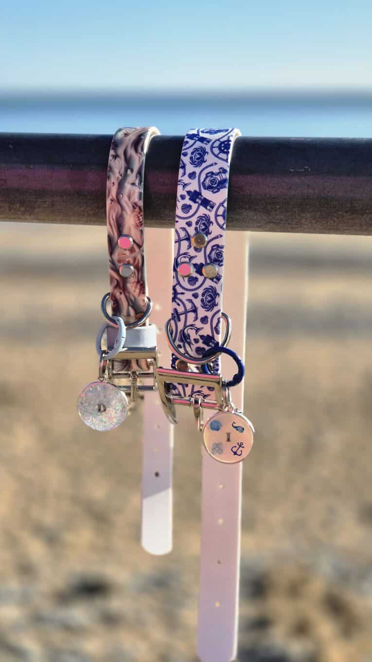 Two coastal-printed BioThane collars with matching personalised resin tags displayed against a beach backdrop.