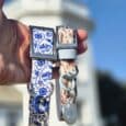 Hand holding coastal-printed BioThane collar with matching personalised resin tag against seaside backdrop.