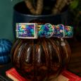 Colourful Halloween-themed handmade dog collar with pumpkin and bat print, displayed on brown glass vessel with books.
